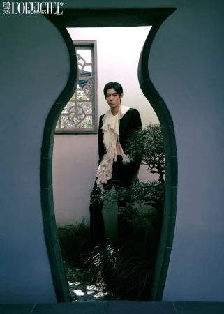 Asian male in a black suit with a ruffled white top framed by a decorative outdoor window surrounded by greenery and garden elements.