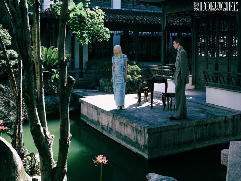 Two people stand on a stone platform surrounded by a pond and traditional Chinese architecture, bathed in soft natural light.