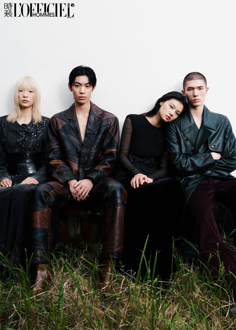 Editorial fashion photo of four Asian models seated together in stylish leather and dark clothing against a white wall background with grassy foreground.