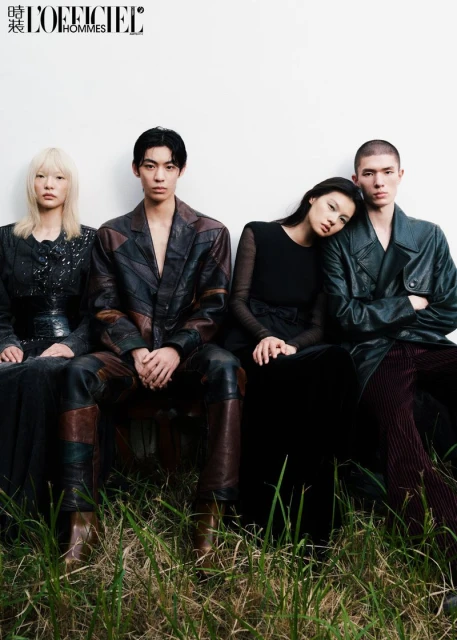 Editorial fashion photo of four Asian models seated together in stylish leather and dark clothing against a white wall background with grassy foreground.