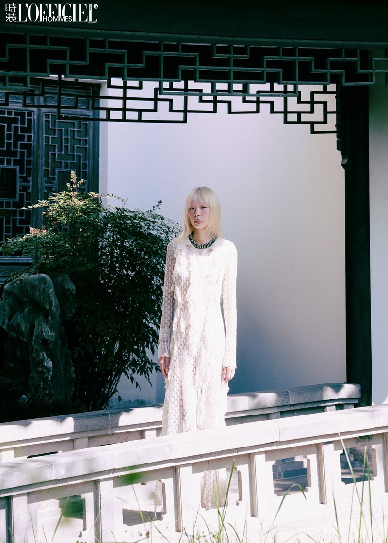 A fair-skinned woman with blonde hair wears a long white lace dress while posing in front of a white wall and dark Asian architecture.