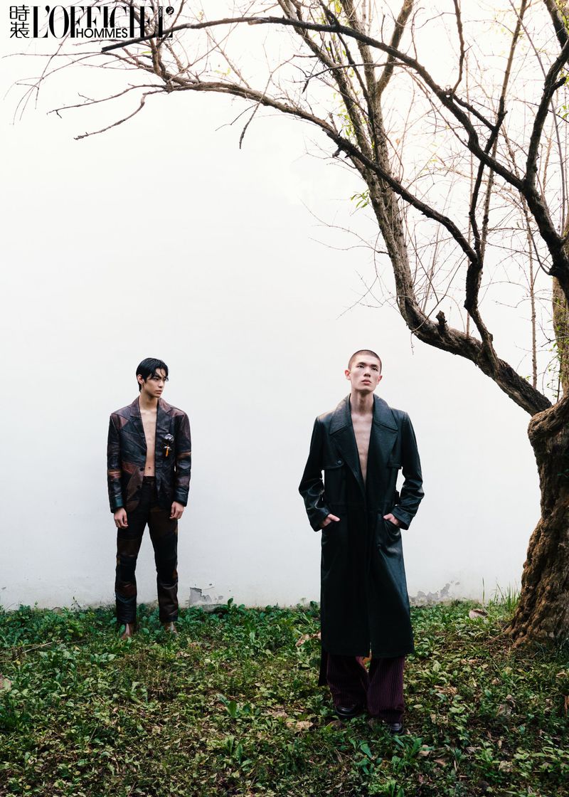 Two male models posing near a bare tree against a white wall with grassy ground for L'Officiel Hommes China editorial