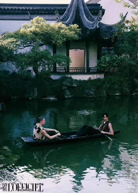 Two people in a boat on a pond with a traditional Chinese building and greenery in the background, editorial fashion photo.