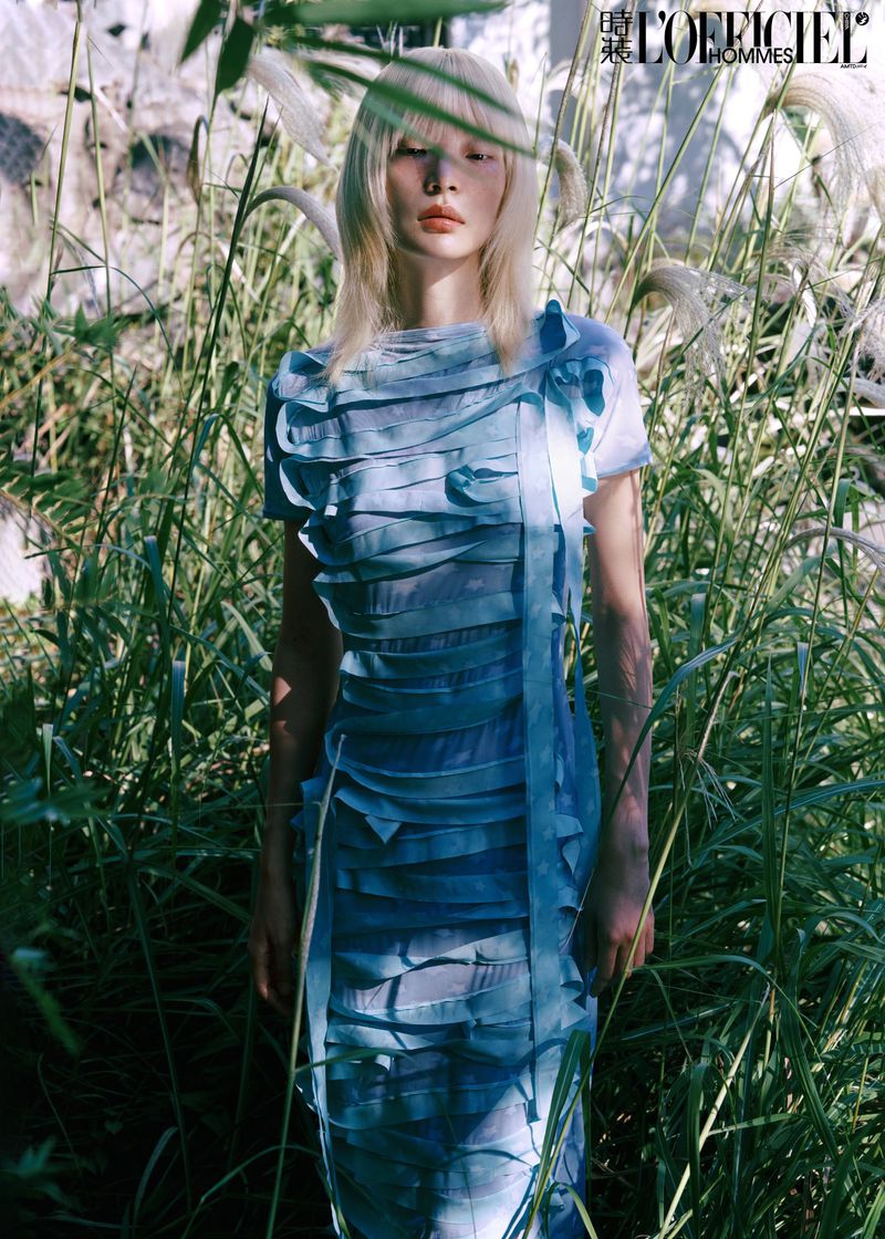 Fashion editorial shot of a female model with platinum blonde hair wearing a light blue layered dress in an overgrown field.