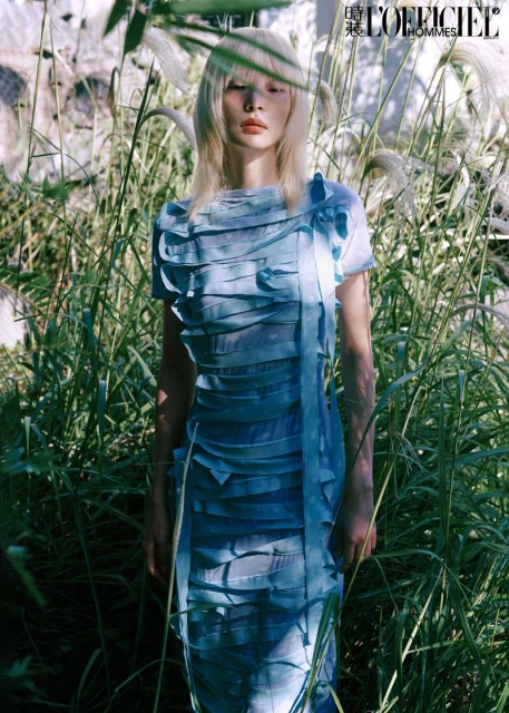 Fashion editorial shot of a female model with platinum blonde hair wearing a light blue layered dress in an overgrown field.