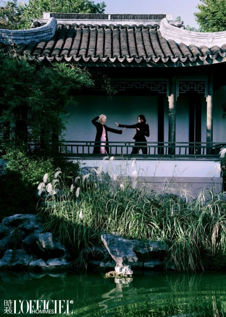 Two people practice martial arts on a veranda with a traditional Chinese roof, overlooking a tranquil garden and pond surrounded by lush foliage.