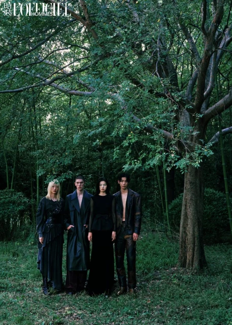 Four fashion models in dark clothing pose in a wooded area with dappled light and tall trees for a dramatic editorial shot.