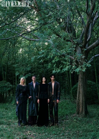 Four fashion models in dark clothing pose in a wooded area with dappled light and tall trees for a dramatic editorial shot.