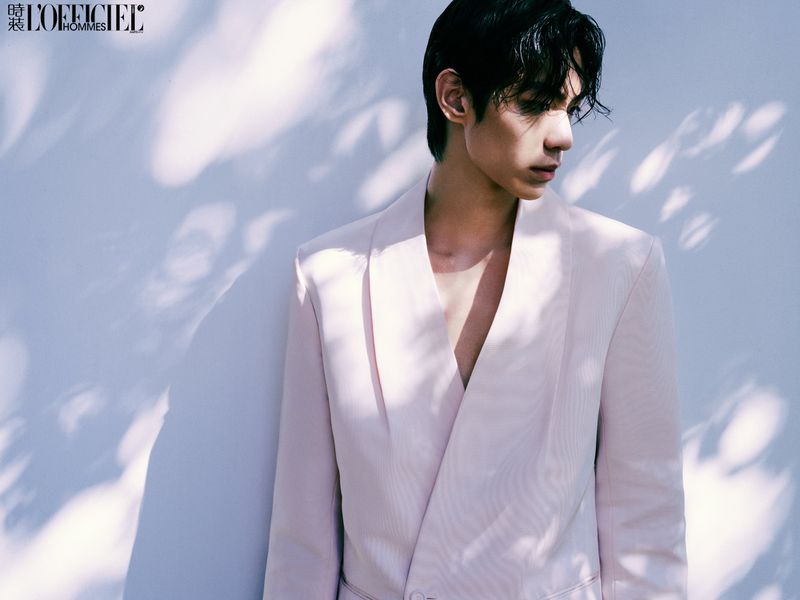 Portrait of a young Asian man in a pink suit, standing in front of a textured light-blue wall with dappled shadows. Fashion editorial.