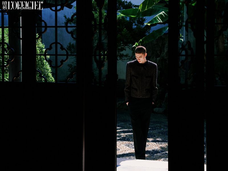 A fashion shot presents an adult man in a patterned jacket and dark pants, walking in a courtyard with lush foliage, framed by an ornate gate.