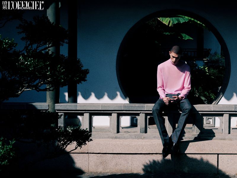Man in pink top sits contemplatively against a round window framed by greenery in an outdoor setting, captured in soft, diffused light.