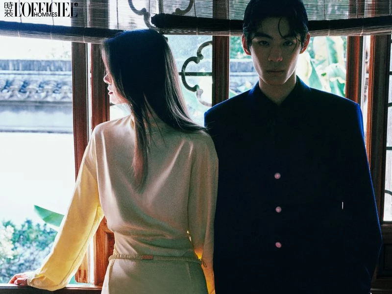 Indoor fashion shot of a male and a female model in a room with natural lighting. The man wears a navy suit and the woman, a light yellow dress.