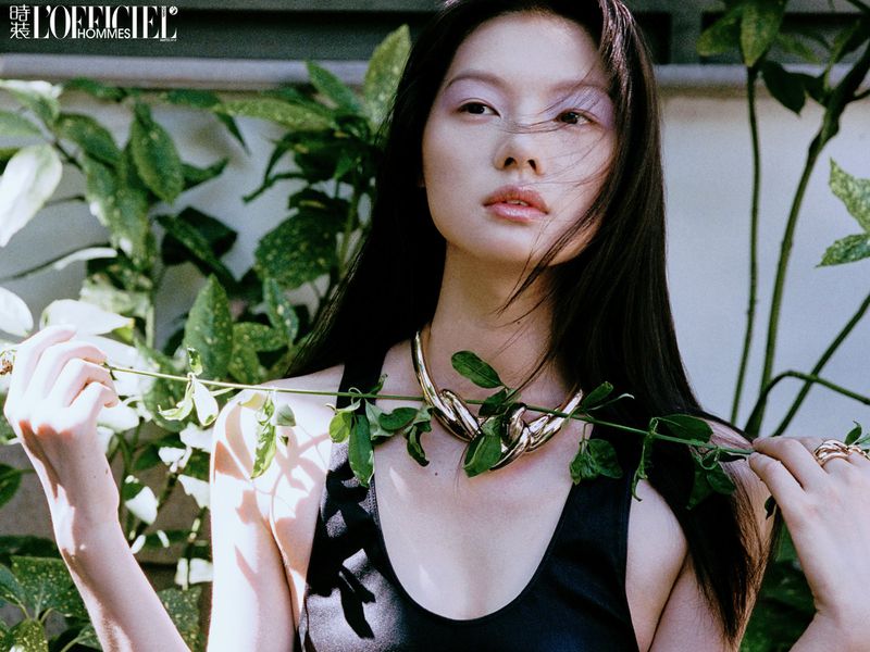East Asian model in a black tank top and sculptural gold necklace holding a green leafy vine against foliage.