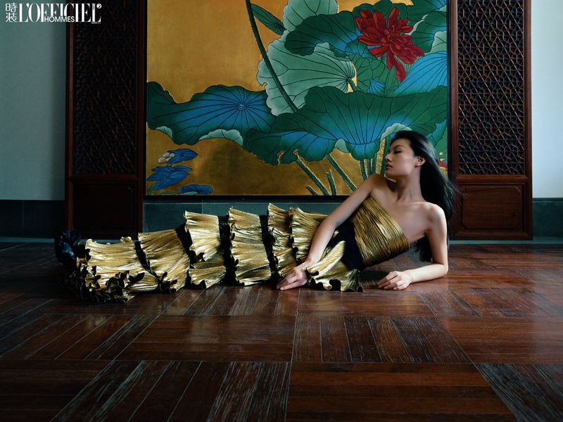 Elegant Asian woman in gold gown lying on a wood floor in front of a gold painting of lotus blossoms for fashion editorial.