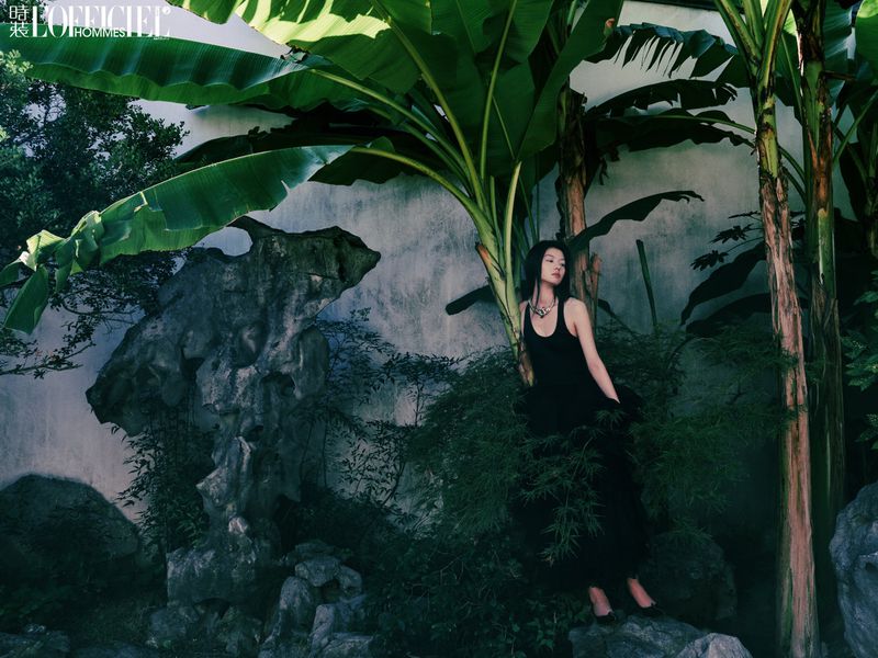 Elegant woman in a black dress poses on rocks surrounded by lush greenery and banana trees in a captivating outdoor fashion shot.