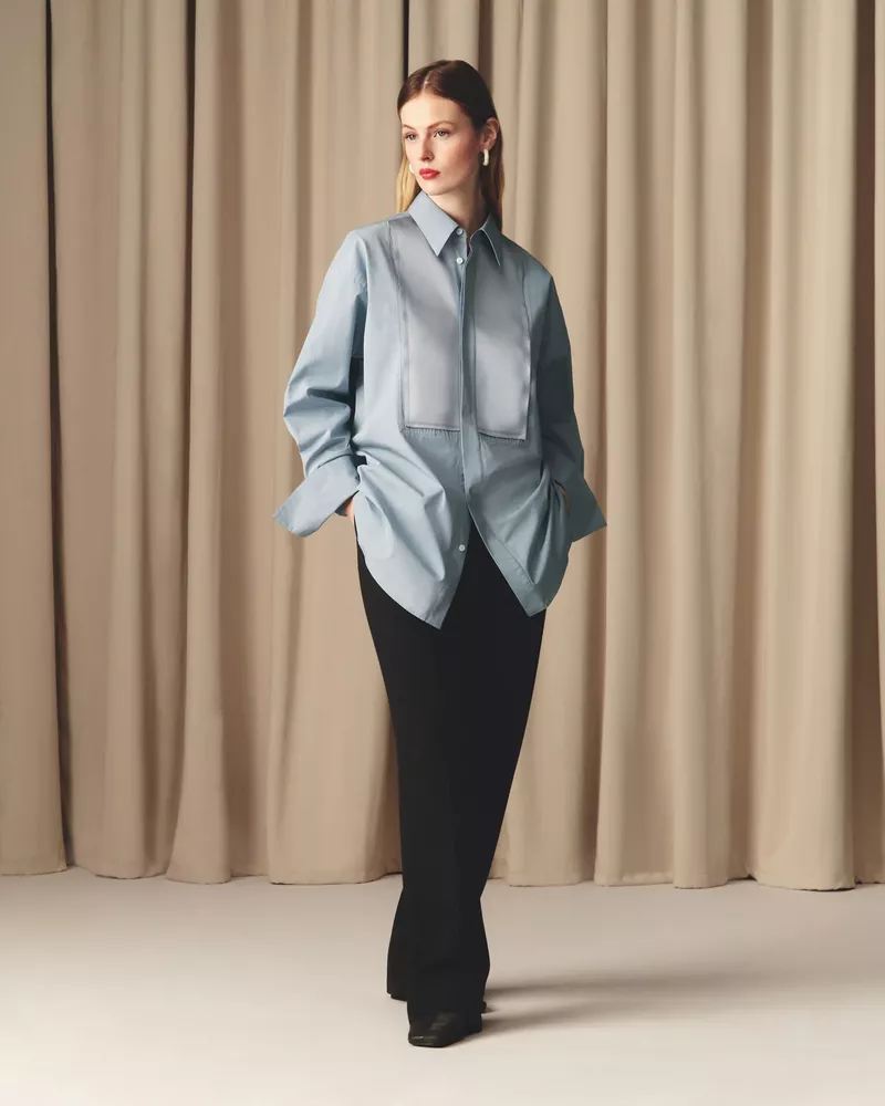 A fashionable woman in a light blue shirt and black pants poses in front of neutral curtains in a studio setting, showcasing a modern style.