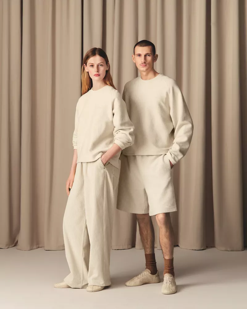 A stylish couple poses in matching neutral loungewear against a soft beige curtain backdrop, showcasing a blend of comfort and fashion.