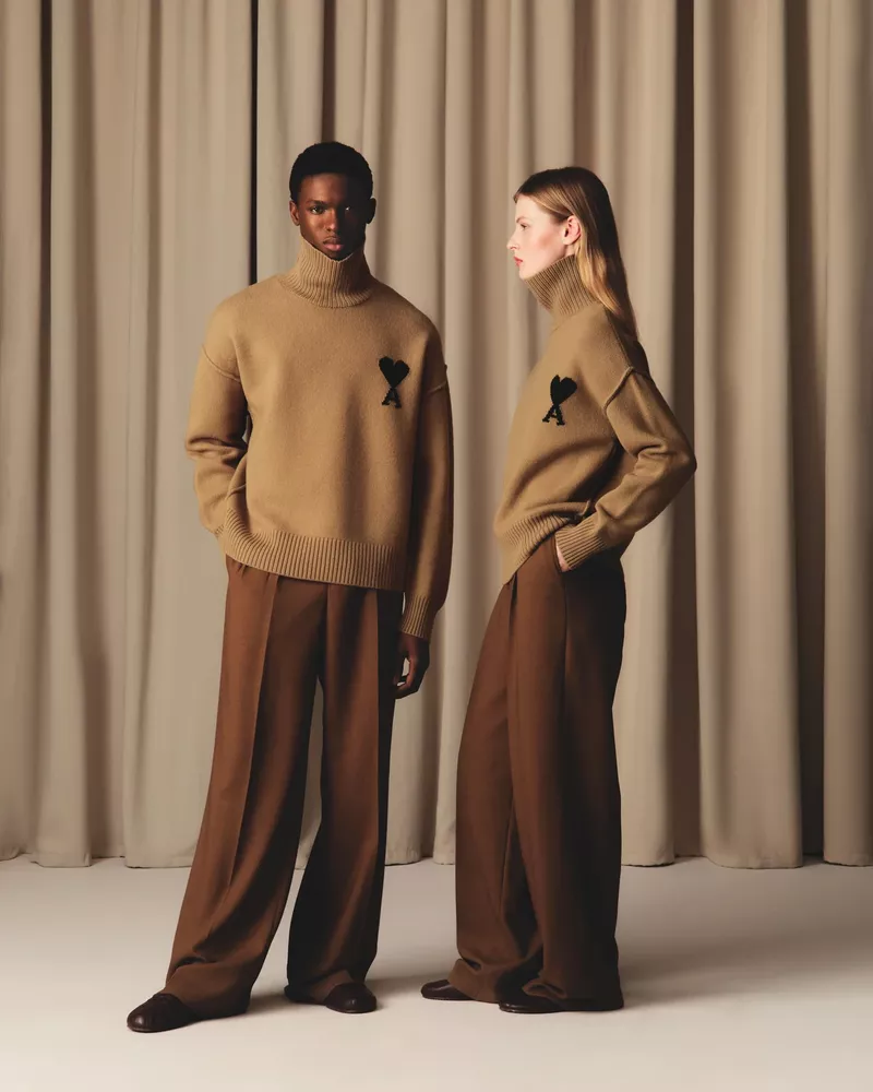 Two fashion models pose wearing matching tan turtlenecks with a heart logo and brown trousers against a neutral curtain backdrop.