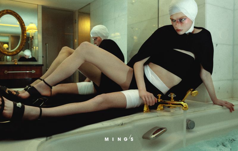 Woman poses in a black outfit and head covering inside a marble-walled bathroom beside a tub with gold fixtures and reflective oval mirror.