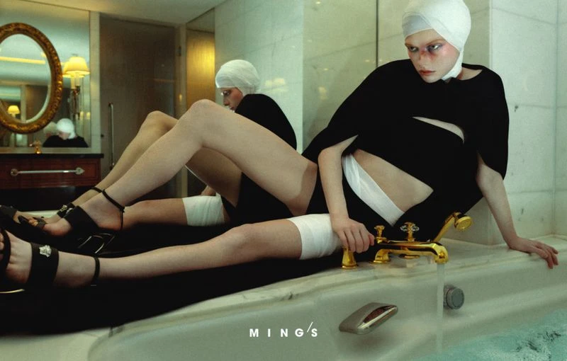 Woman poses in a black outfit and head covering inside a marble-walled bathroom beside a tub with gold fixtures and reflective oval mirror.