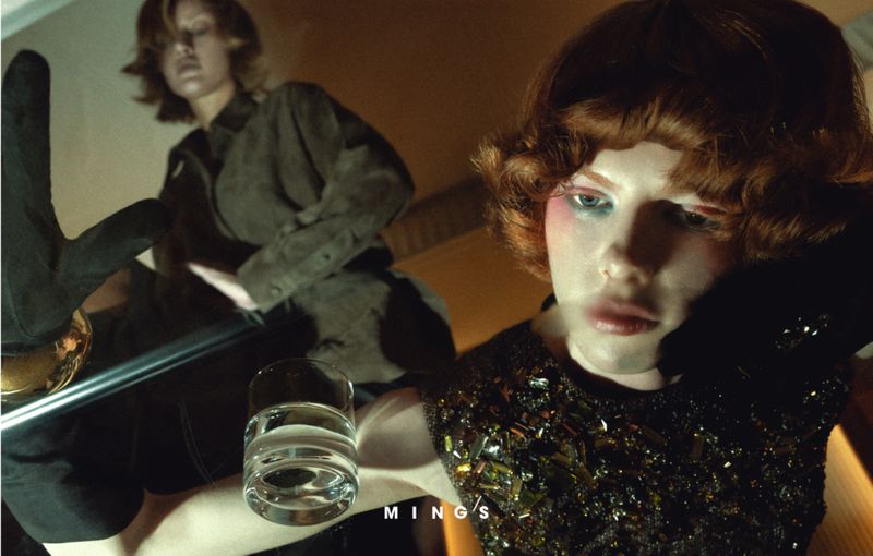Two young adult women in a dimly lit interior; one sits facing away, the other faces forward wearing dark clothing with light makeup.