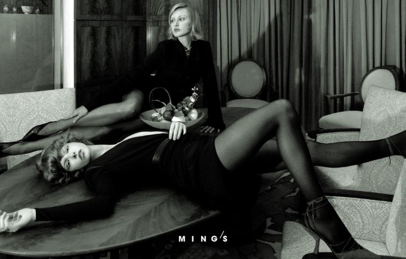 Black and white image features two adult women in a moody indoor setting with armchairs and a round table.
