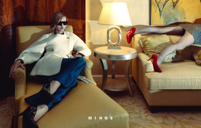 Model wearing dark sunglasses, white jacket and blue trousers seated in hotel room with another figure lounging