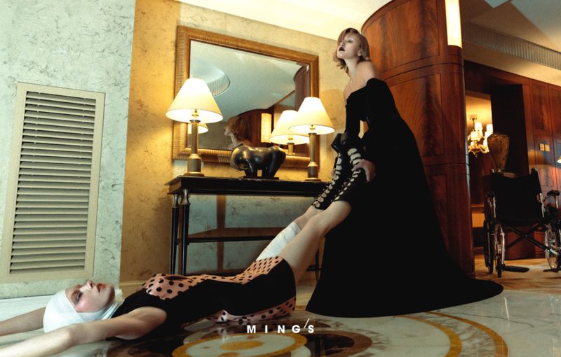 Fashion shot in a hallway with two women: one standing in a black dress and the other lying on the floor wearing a polka-dot dress.