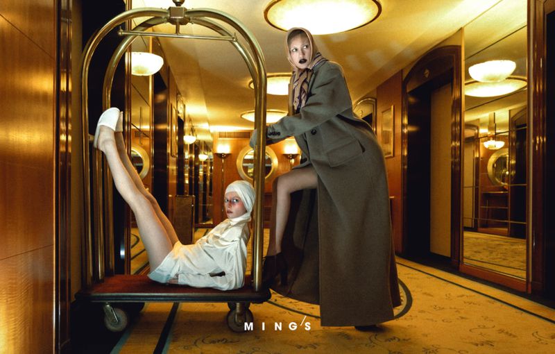 Two women pose with luggage cart in a vintage hotel hallway, one sitting on the cart with legs up, the other pushing it, in editorial fashion.