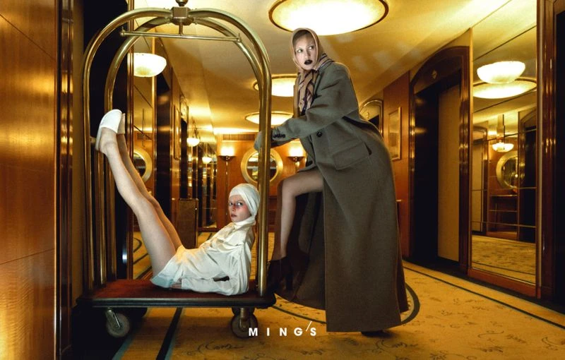 Two women pose with luggage cart in a vintage hotel hallway, one sitting on the cart with legs up, the other pushing it, in editorial fashion.