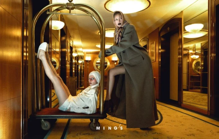 Two women pose with luggage cart in a vintage hotel hallway, one sitting on the cart with legs up, the other pushing it, in editorial fashion.