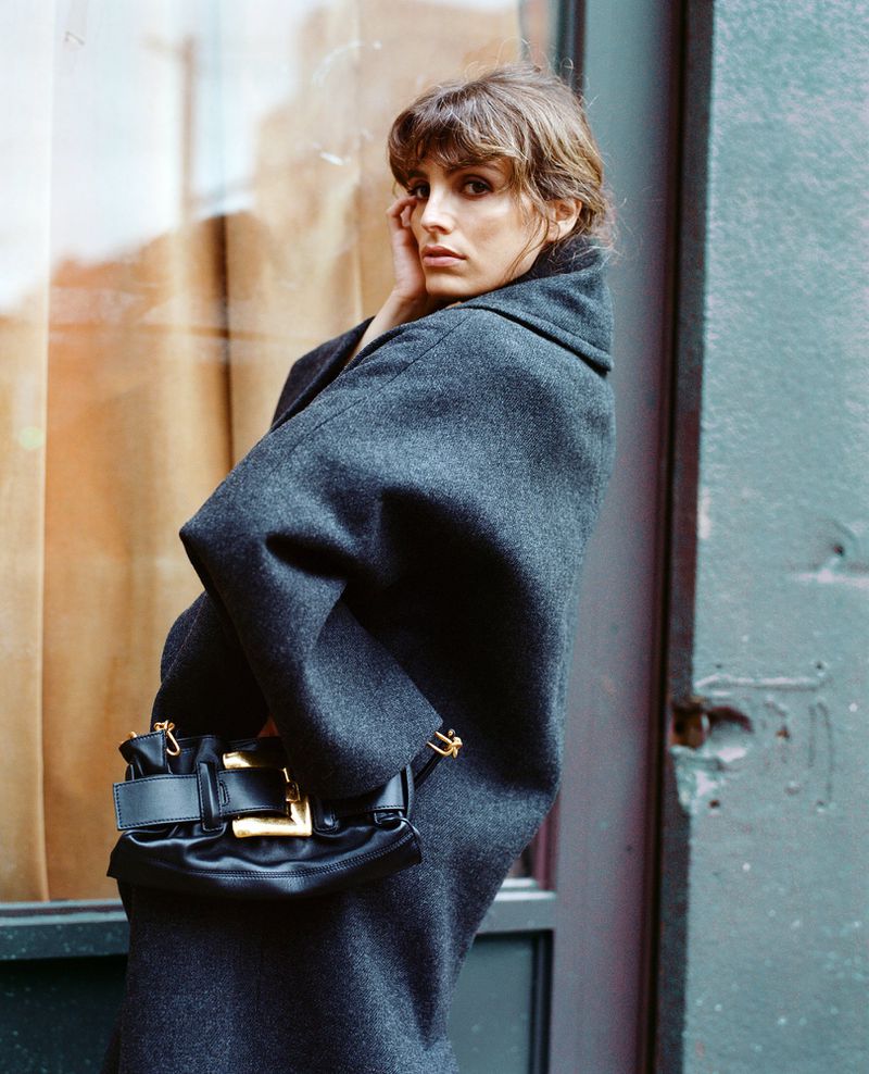 A fashionable adult female model poses in a grey wool coat with a black leather purse with gold hardware in an urban setting.