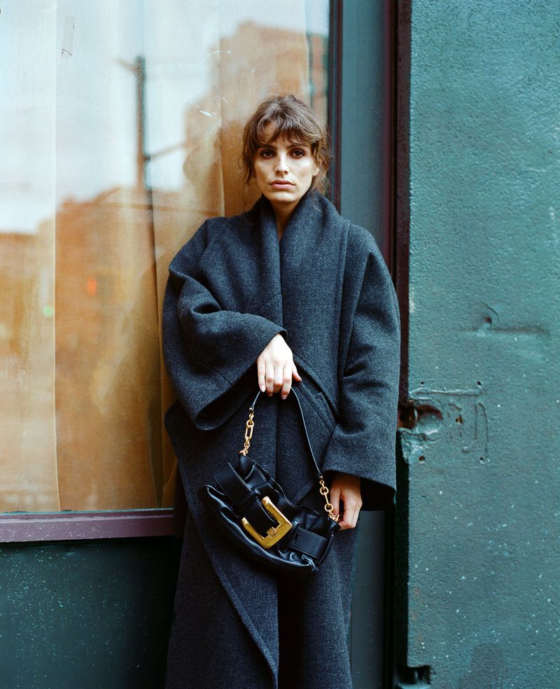 Woman wearing a long wool coat holds a black purse with a gold buckle in front of a window and green painted wall outdoors.