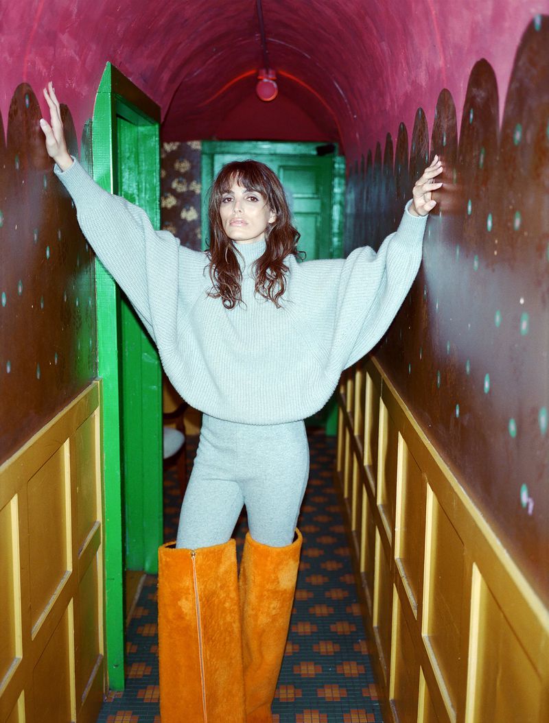 A woman in a sweater and orange boots poses in a hallway with a patterned wallpaper and ceiling in an indoor setting with overhead lighting.