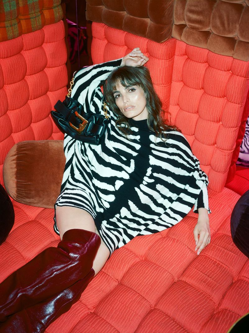 Stylish adult female poses indoors with a zebra-patterned dress, thigh-high brown boots, and black purse, lying on an orange quilted sofa.