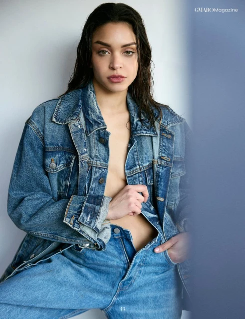 Attractive young woman in denim jacket and jeans poses against a white wall with blue accent. Wet hair and natural makeup.