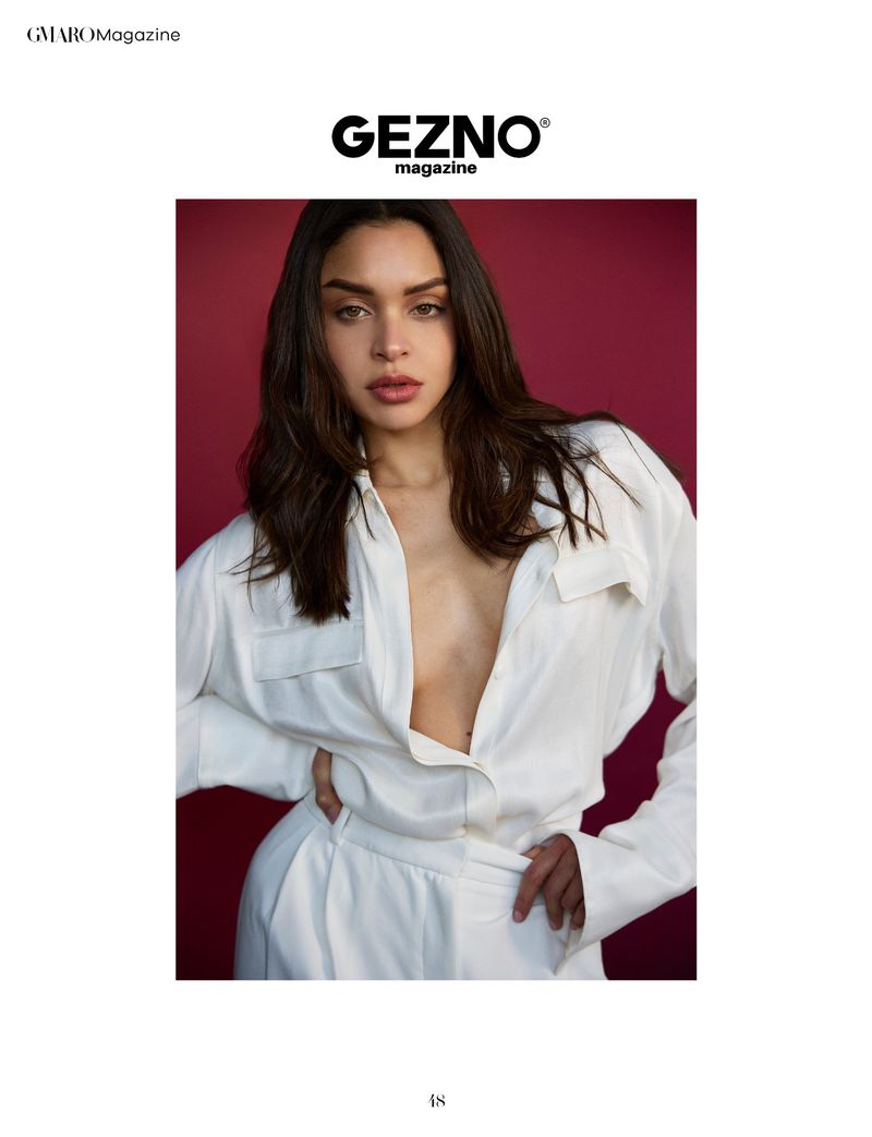Close-up studio portrait of a brunette adult female fashion model in a white oversized shirt, posing against a dark red backdrop.