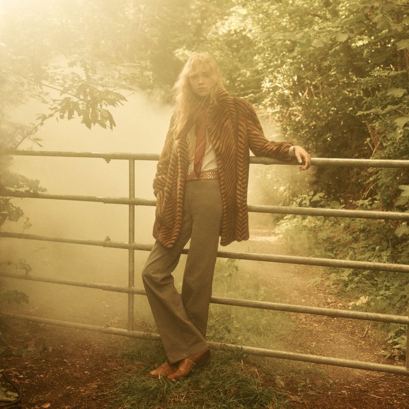 A blond adult standing by a metal fence in a natural, wooded outdoor setting, wearing a striped coat and tie, in a fashion editorial style.