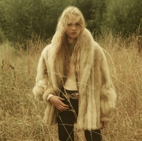 Young woman with long blonde hair wearing a fur coat in a field of tall golden grass, with trees visible in the background.