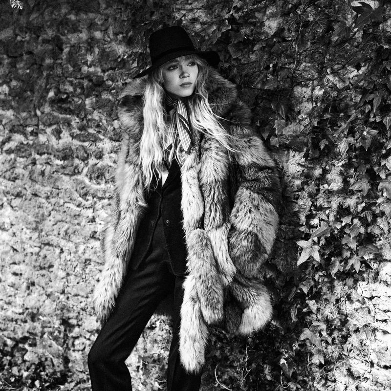 Model Ella Dalton wearing a fur coat and wide-brim hat against a stone wall for Mastermind Magazine.