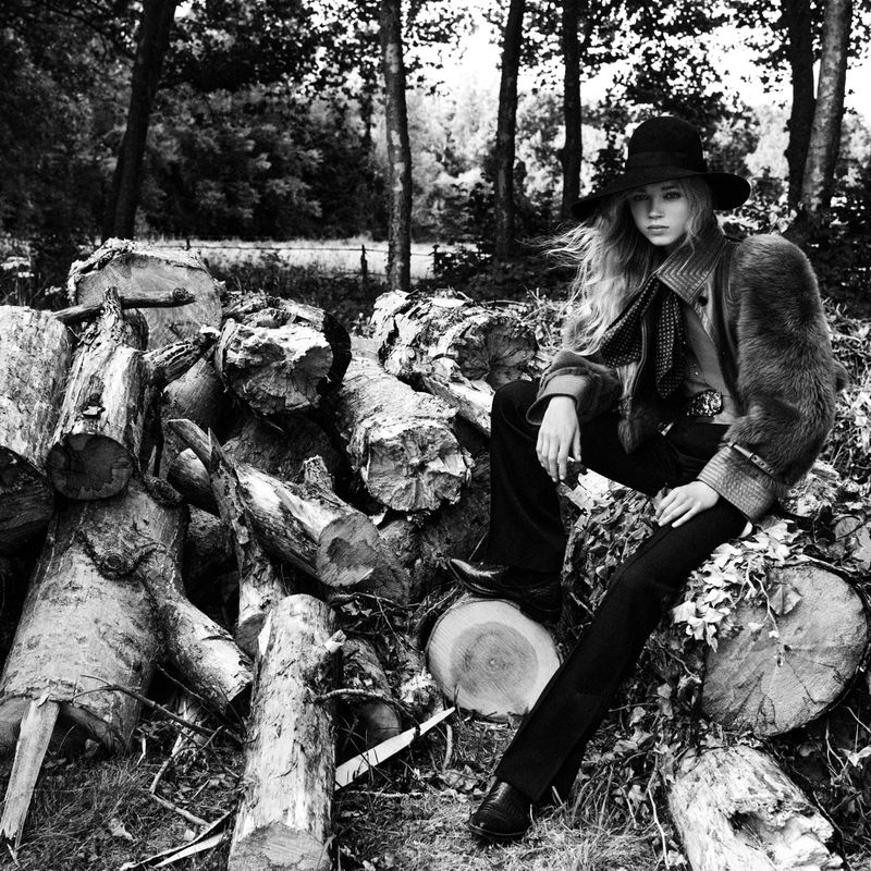 A black and white portrait of a model in a hat and faux fur coat, seated on a pile of logs in a forest setting.