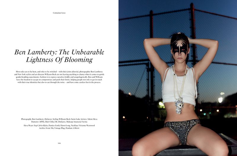 A fashion shot of a young woman in dark briefs and a light tank top is sitting with her arms up against a chainlink fence at night.