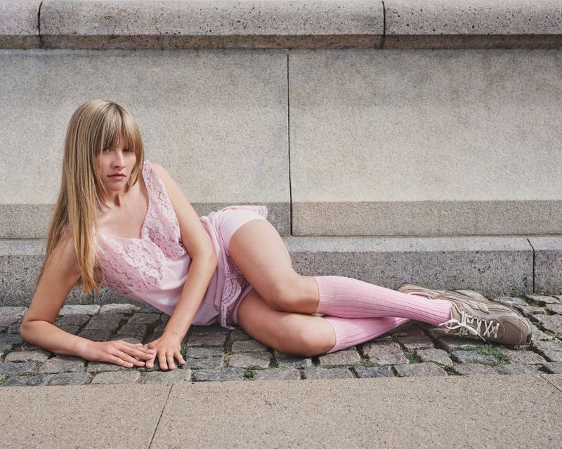 Blonde woman in pink lace lingerie, socks, and sneakers lies on cobblestone in front of a textured concrete wall in an urban setting.