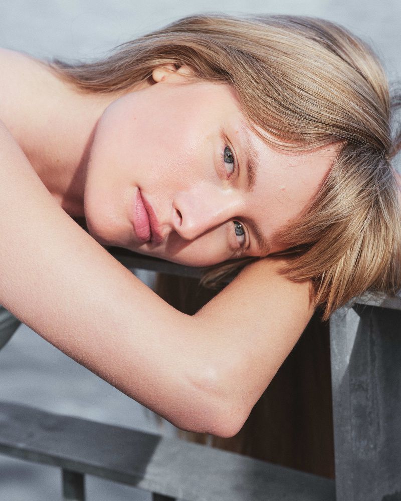 Close-up portrait of a blonde adult female lying down with her arm resting on a grey metal surface, gazing at the camera in a photo studio.