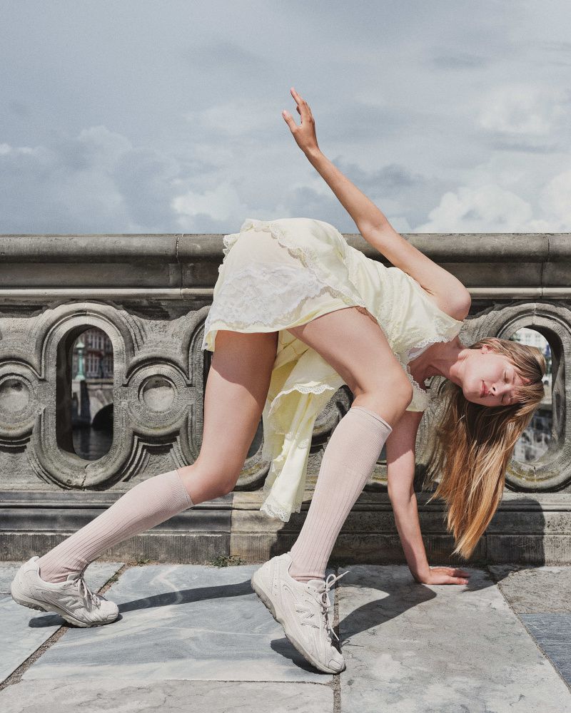 Woman in yellow dress and white sneakers performs acrobatic pose near stone bridge, under a cloudy sky. Fashion and fitness editorial.