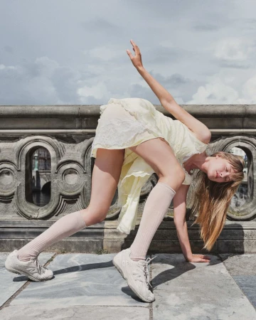 Woman in yellow dress and white sneakers performs acrobatic pose near stone bridge, under a cloudy sky. Fashion and fitness editorial.