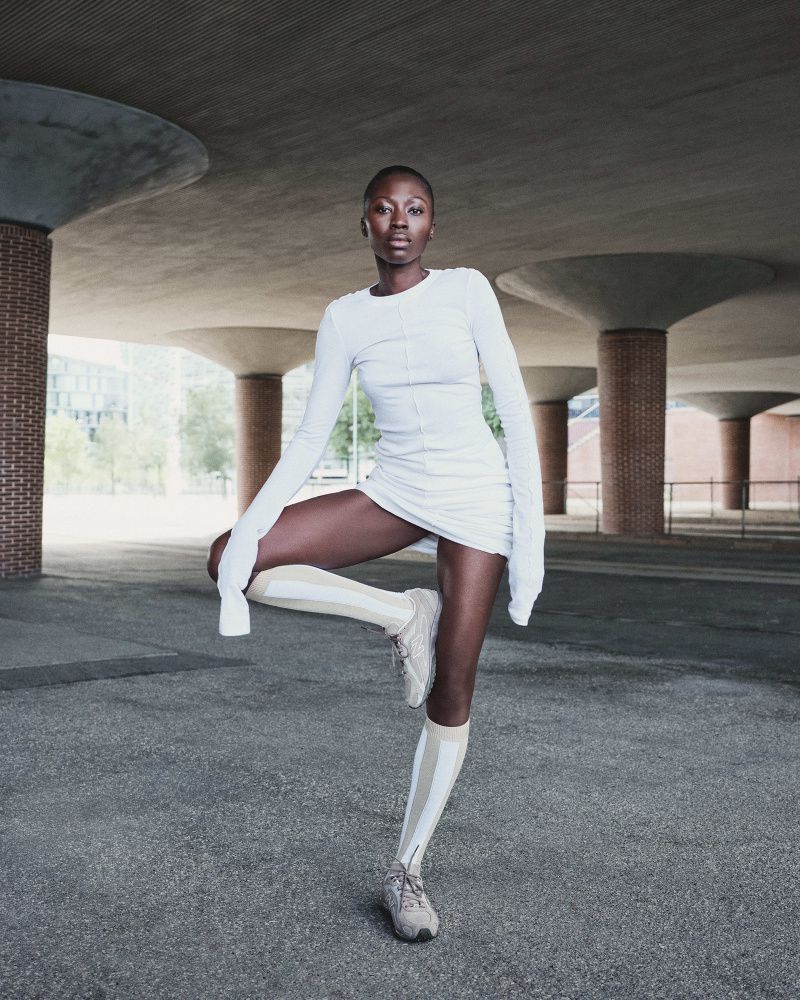 Adult female in a light dress and striped socks stands in a concrete urban environment. The shot emphasizes minimalist fashion and modern architecture.