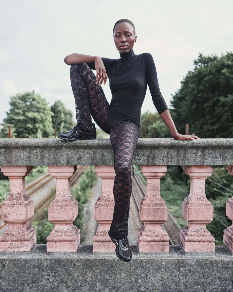 An adult woman in a black turtleneck and patterned tights sitting on a balustrade in an urban park setting, with trees and sky in the background.