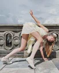 Model in a pale yellow dress and sneakers poses dynamically on a stone bridge against an overcast sky.