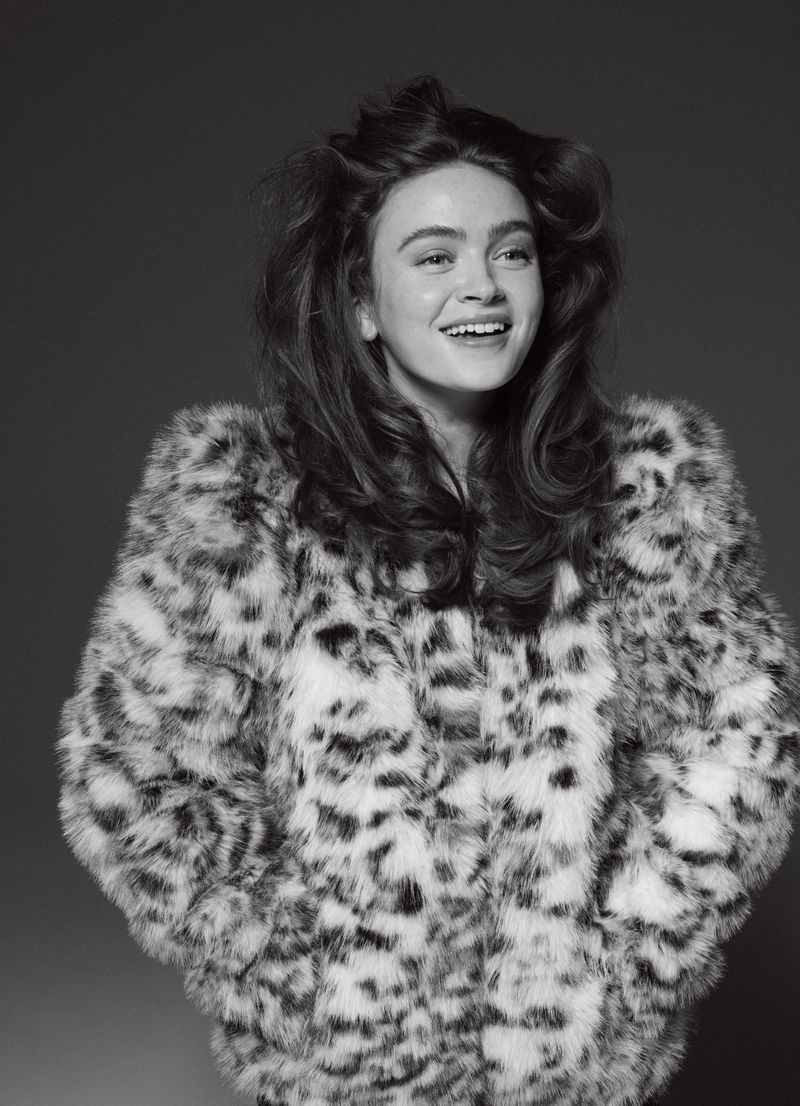 Portrait of a smiling young woman with long wavy brown hair wearing a patterned faux fur coat in a studio setting, captured in black and white.